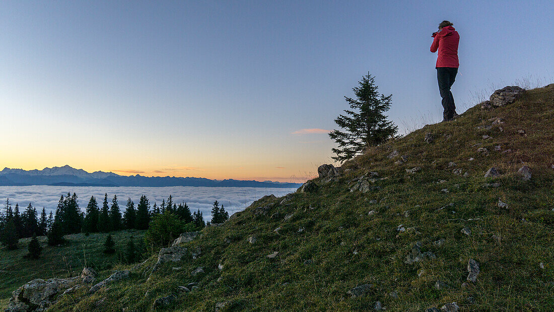 someone with red coat and black pants photographing the sunrise over the sea of clouds on the Geneva Lake with the Alpine chain in the background, in the Vaud Canton, Switzerland