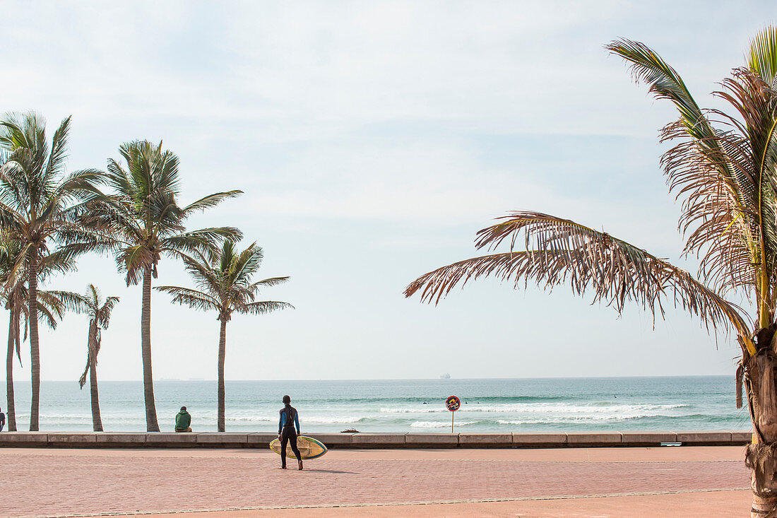 Ein Junge mit einem Surfbrett geht die Promenade entlang an der Goldenen Meile - benannt nach den goldenen Sandstränden - Durban, Südafrika.