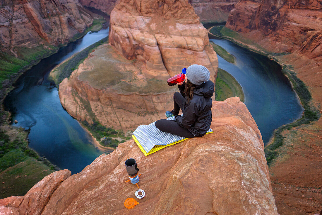 Female hiker sitting and drinking coffee on edge of cliff at Horseshoe Bend of Colorado River, Arizona, USA