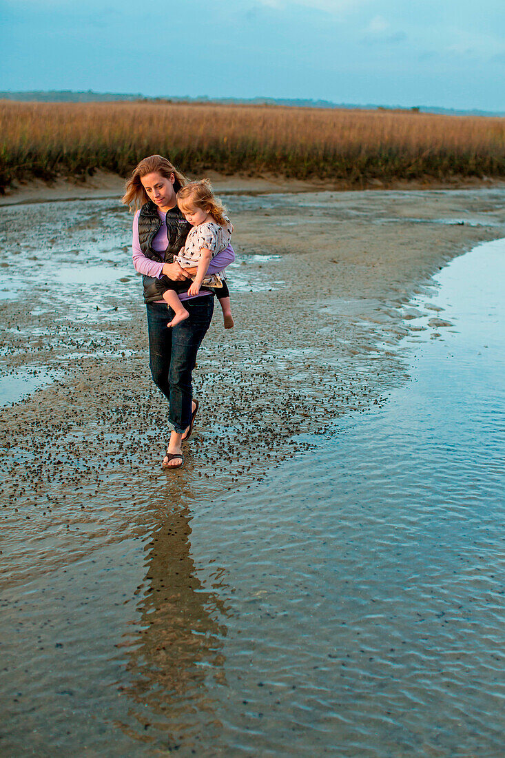 A woman walks near shallow water carrying her daughter in her arms in a marsh at Wrightsville Beach, North Carolina.