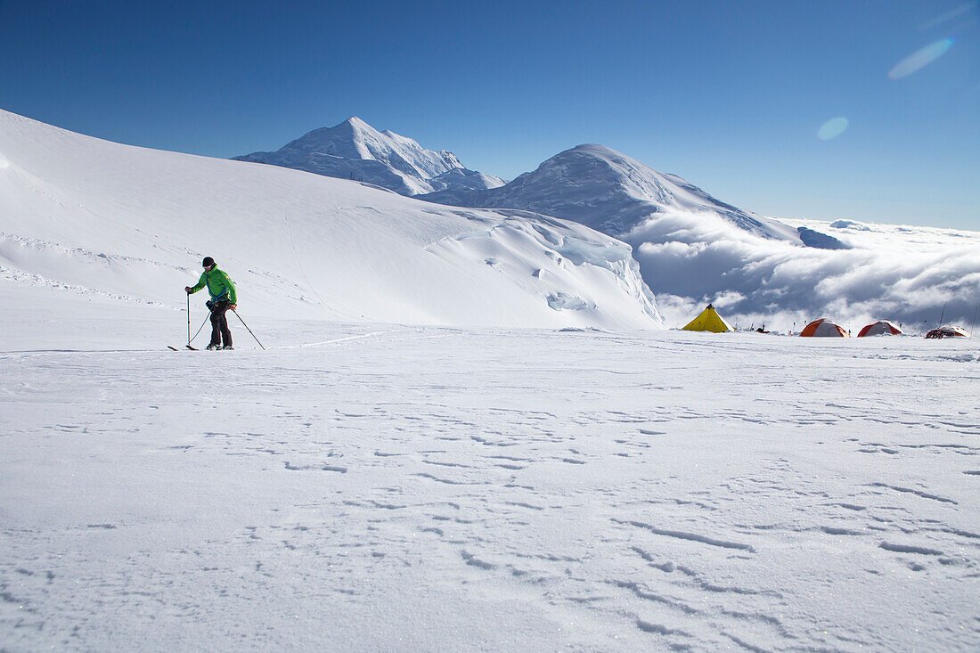Ein Ski-Bergsteiger auf dem Kahiltna-Gletscher des Denali-Nationalparks in Alaska, mit Mount Foraker und Kahiltna Dome im Hintergrund. Auf der rechten Seite sind die Zelte von 11 K Camp auf Denali sichtbar.