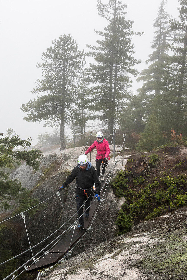A man and a women cross a small suspension bridge that is part of the Via Ferrata in Squamish, British Columbia.