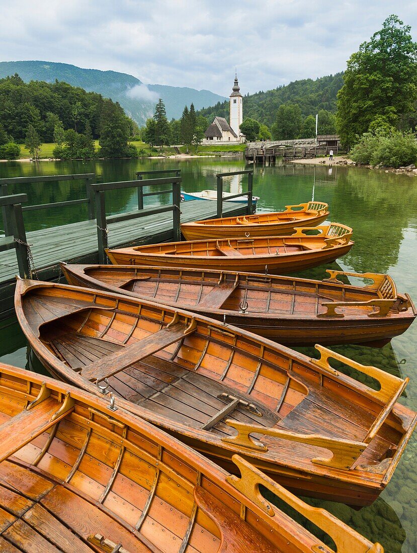 Bohinjer See (Bohinjsko jezero), Triglav-Nationalpark, Oberkrain, Slowenien. Ruderboote zu vermieten. Die Kirche von St. John (Cerkev SV Janeza) in Ribcev Laz im Hintergrund.
