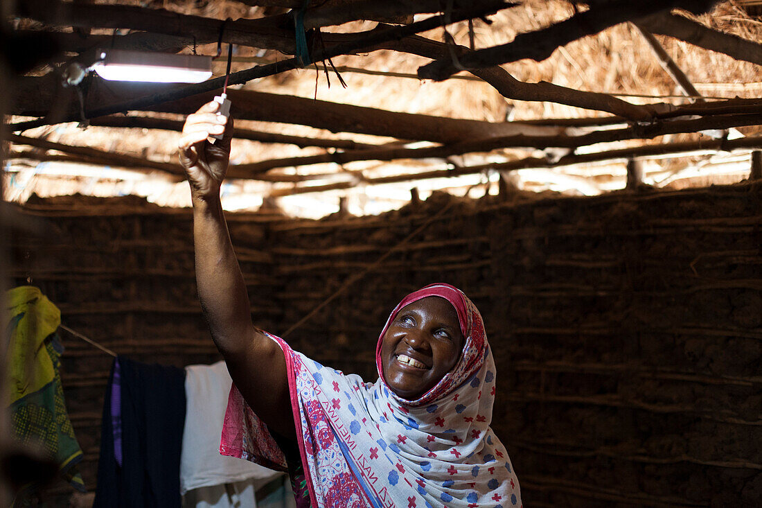 Eine Frau lächelt, als sie das neue Sonnenlicht in ihrer Lehmhütte einschaltet, Tansania, Ostafrika, Afrika