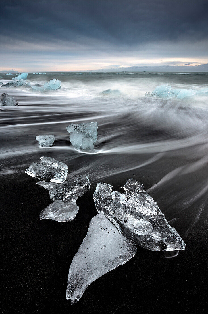 Gläserne Stücke von Eis auf vulkanischen schwarzen Sandstrand mit verschwommenen Wellen, in der Nähe von Jokulsarlon Lagune, Südisland, Polargebiete