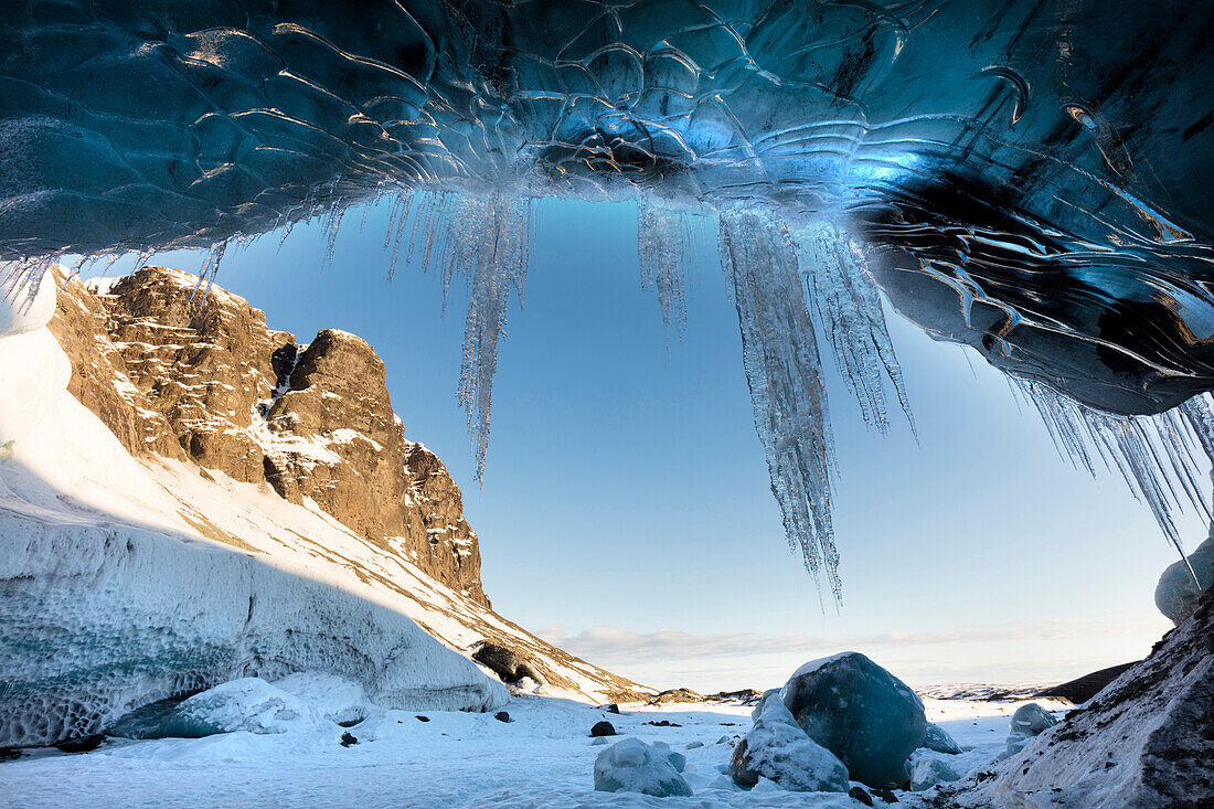 Blick von der Eishöhle auf sonnenbeschienene Berge mit Eiszapfen am Höhleneingang, nahe Jokulsarlon, Süd-Icelan, Polargebiete