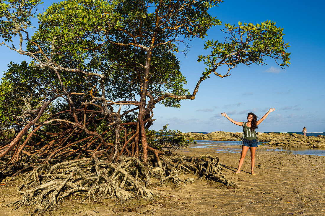 Photograph of young woman with open arms standing near mangrove trees on tropical beach, Morro de Sao Paulo, Bahia, Brazil