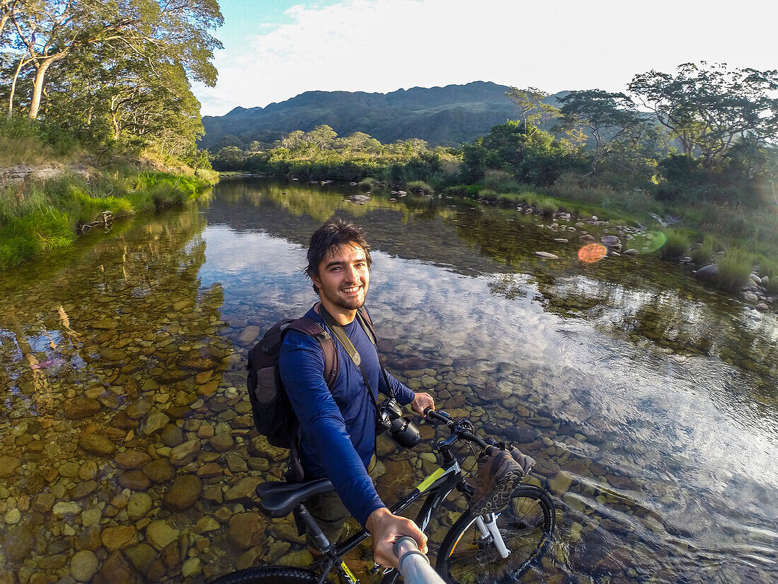 Photograph of smiling man with bicycle taking selfie while crossing Mascates River in Serra do Cipo National Park, Minas Gerais, Brazil