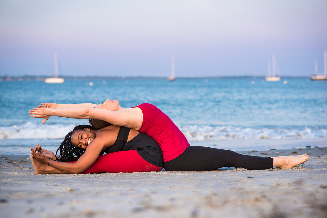Zwei Frauen machen Yoga am Strand in Assisted Fish Pose Variation (Matsyasana) und Seated Forward Fold Pose (Uttanasana), Newport, Rhode Island, USA
