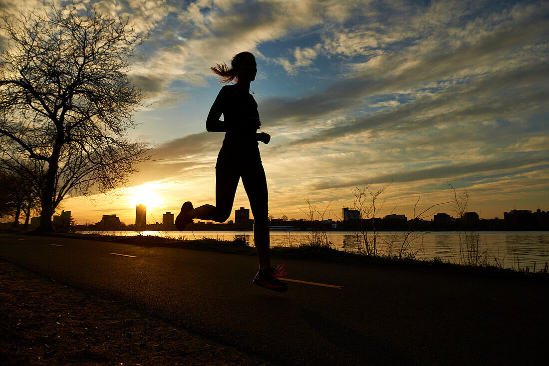 Silhouette of woman jogging on riverside road at cloudy sunset, Boston, Massachusetts, USA