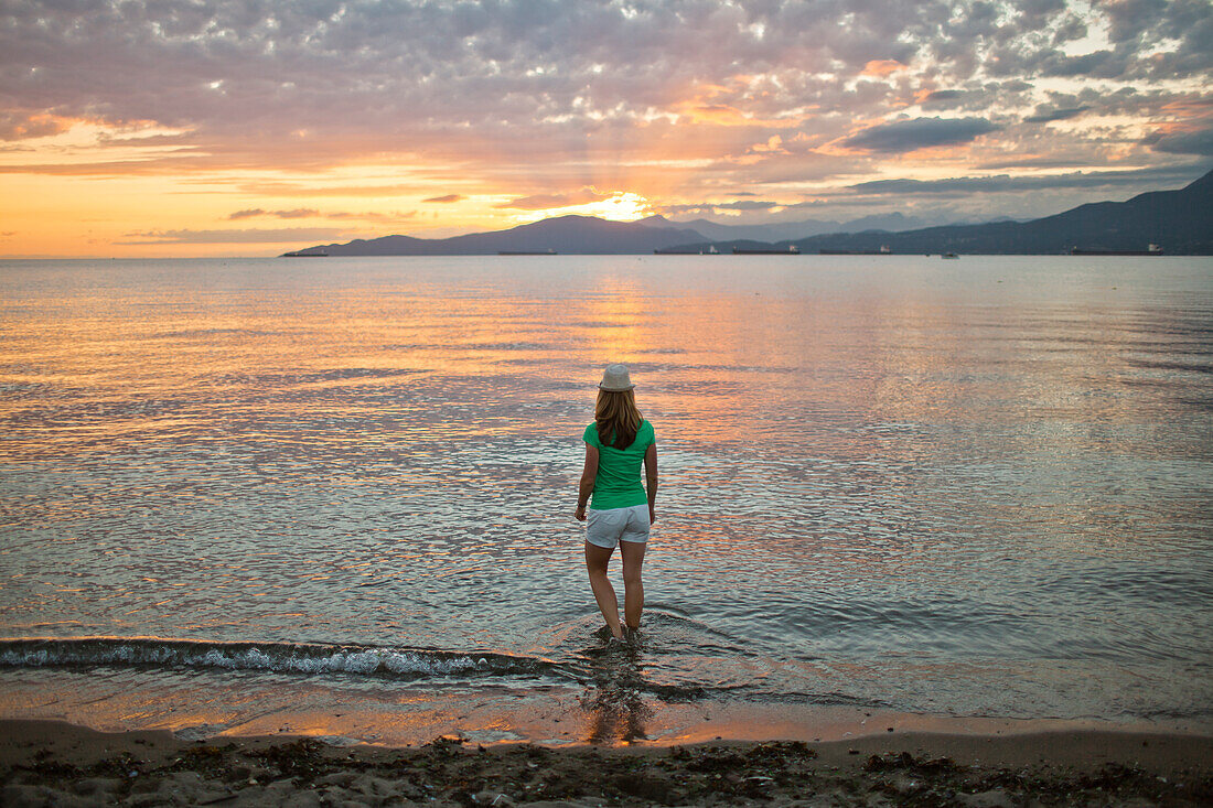 Rear view photograph of woman standing ankle deep in water at Kitsilano Beach during sunset, Vancouver, British Columbia, Canada