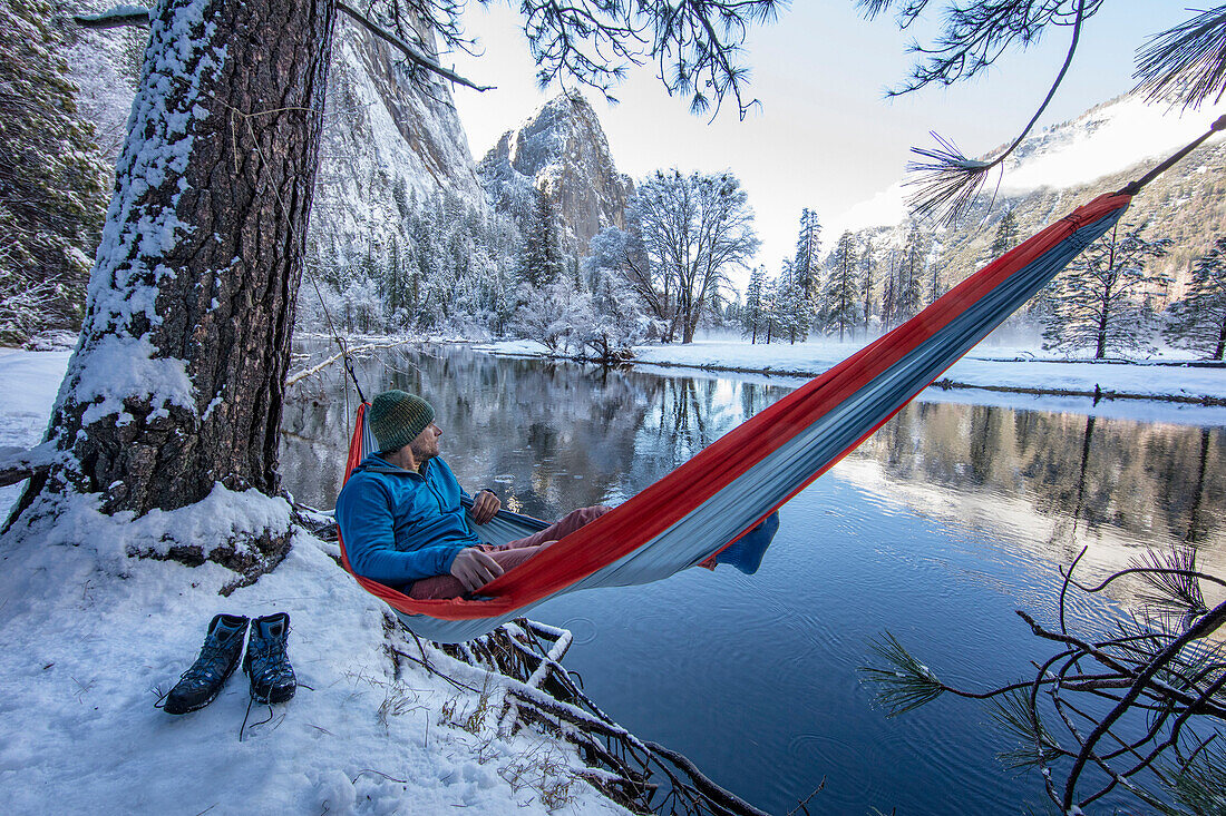 Photograph of man resting in hammock on lakeshore during winter hiking, High Sierras, California, USA