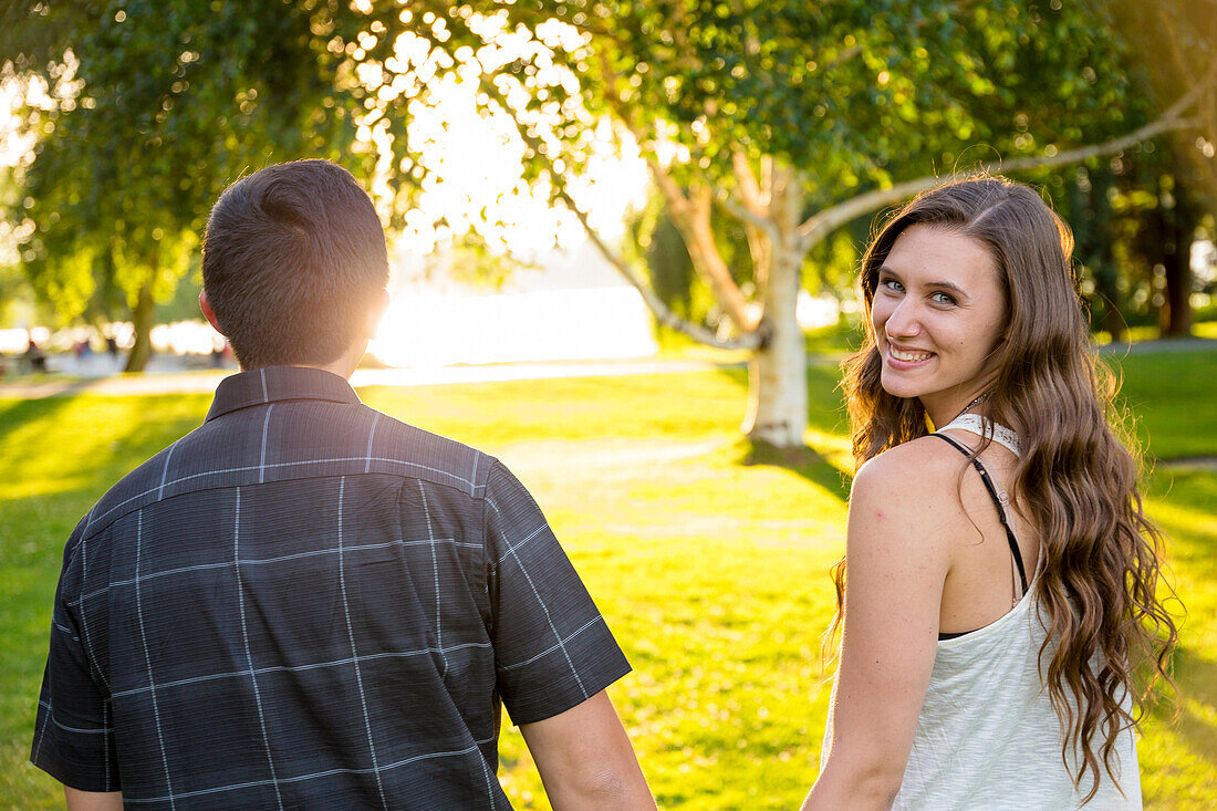 Rear view photograph of couple walking holding hands and woman looking back at camera, Green Lake Park, Seattle, Washington State, USA