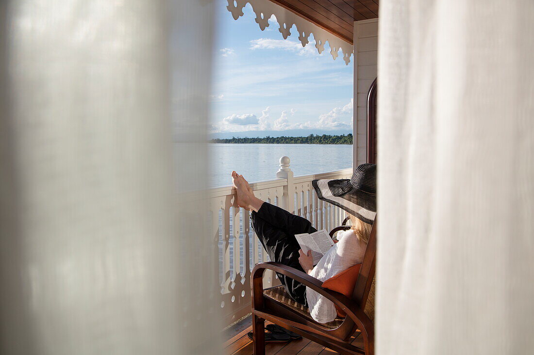 Young woman relaxes on balcony of Executive Suite aboard Ayeyarwady (Irrawaddy) river cruise ship Anawrahta (Heritage Line), near Kyunttaw, Kachin, Myanmar