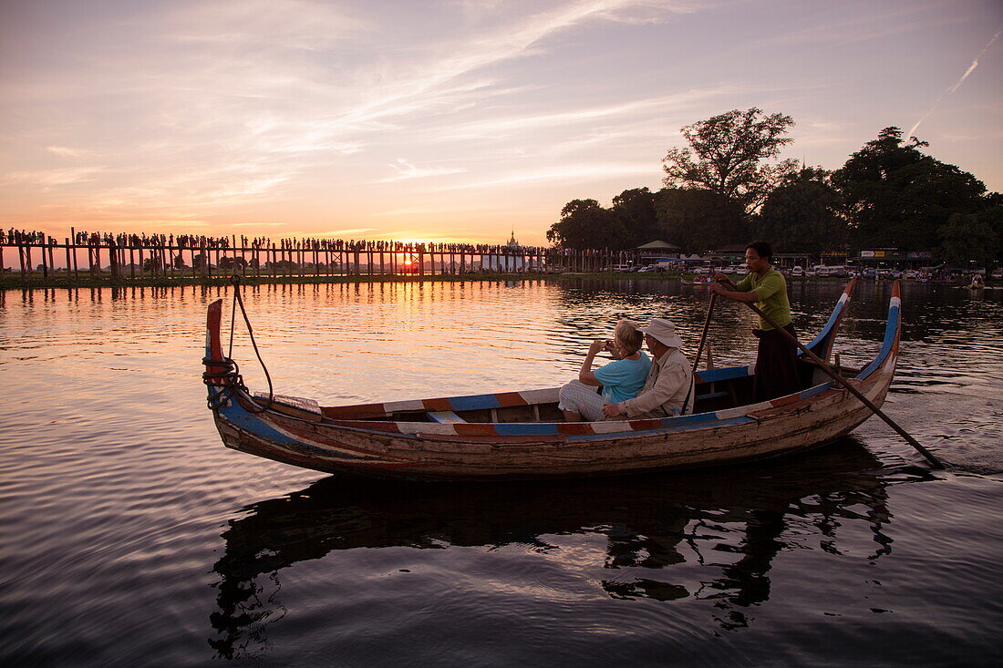 Tourists on row boat with silhouette of people walking along U Bein Bridge across Taungthaman Lake at sunset, Amarapura, Mandalay, Myanmar