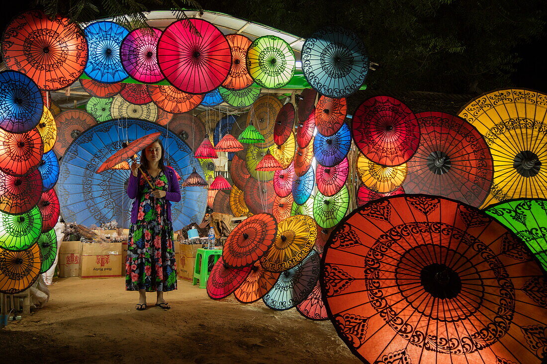Woman holds umbrella in shop offering colorful umbrellas at night, Nyaung-U, near Bagan, Mandalay, Myanmar