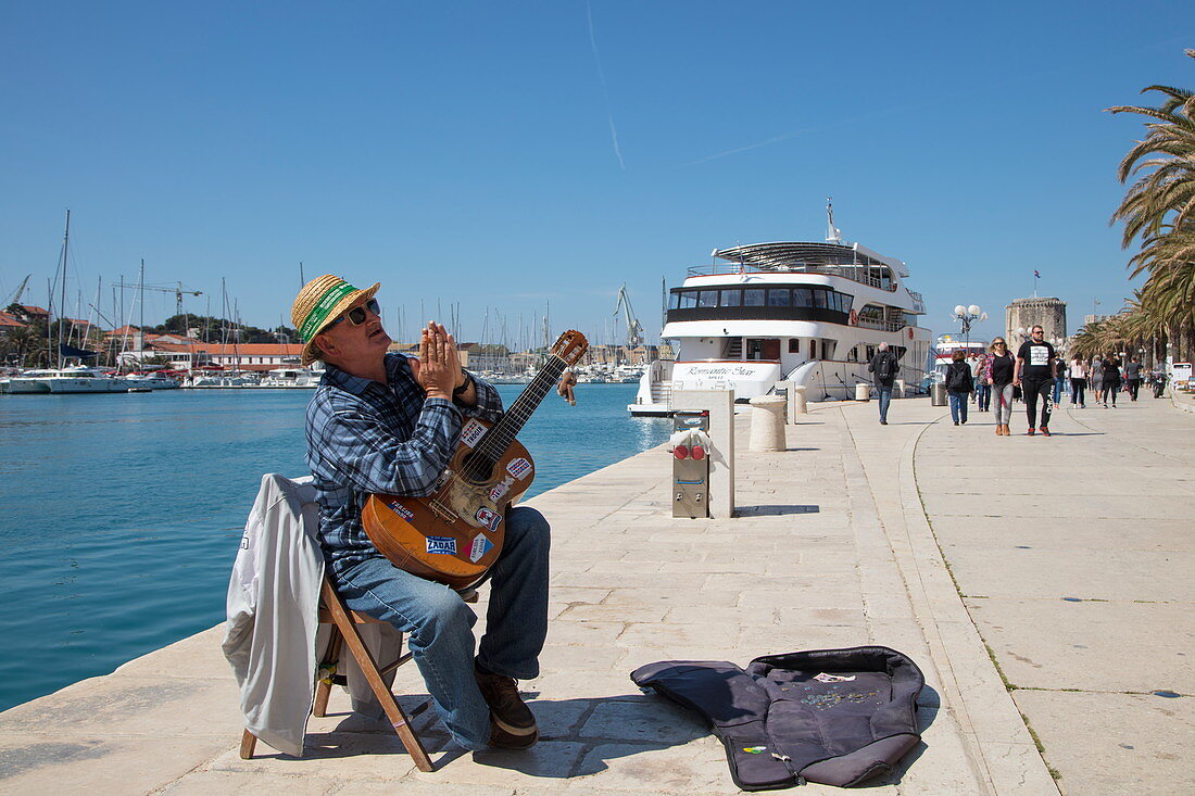 Street musician plays guitar and performs along seafront promenade with cruise ship MS Romantic Star (Reisebüro Mittelthurgau) behind, Trogir, Split-Dalmatia, Croatia