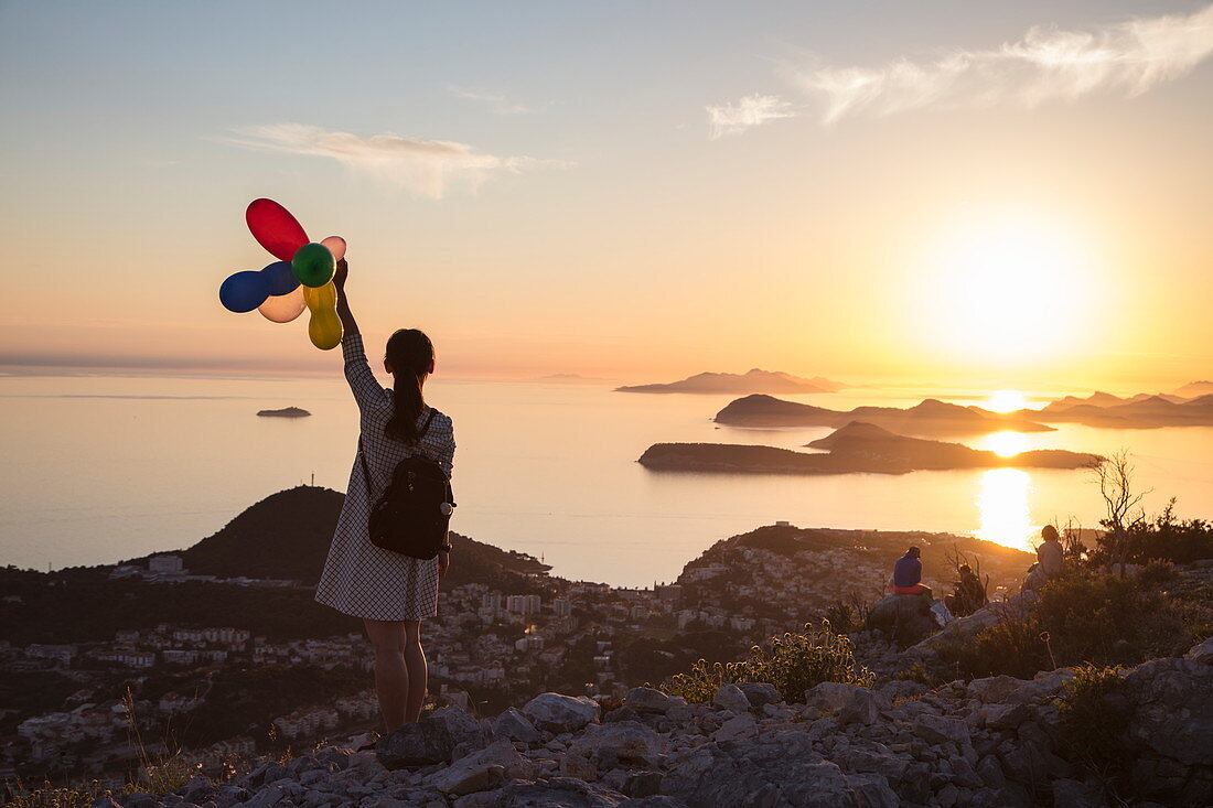 Junge Asiatin hält bunte Luftballons und genießt Aussicht über die Lapad-Halbinsel und vorgelagerte Inseln, vom Hügel nahe der Bergstatation der Dubrovnik-Gondel bei Sonnenuntergang aus gesehen, Dubrovnik, Dubrovnik-Neretva, Kroatien, Europa