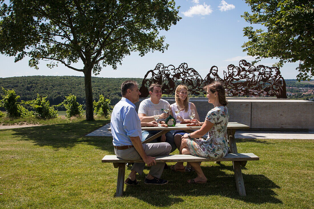'People sit at picnic table in front of ''Wein & Bibel'' sculpture (modelled after The Last Supper) at Terroir F site at Kapellenberg vineyard, Frickenhausen, near Ochsenfurt, Franconia, Bavaria, Germany'