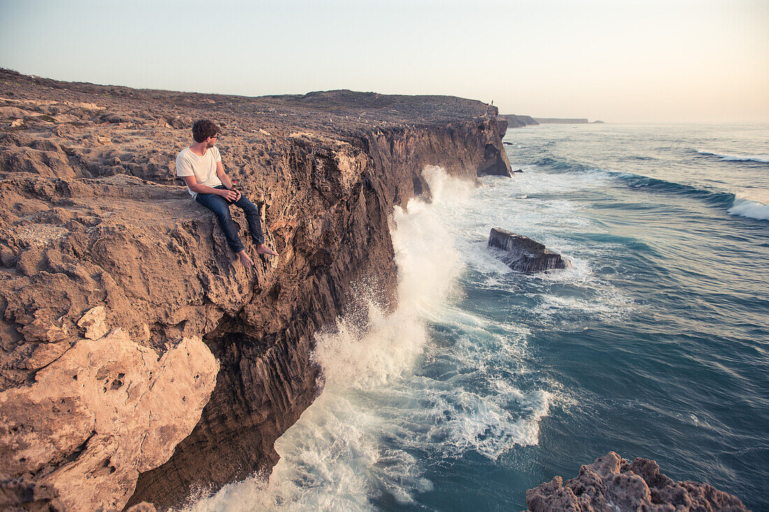 Young man sitting on a cliff edge at the sea at the beach Praia da Amoreira,  Aljezur, Faro, Portugal