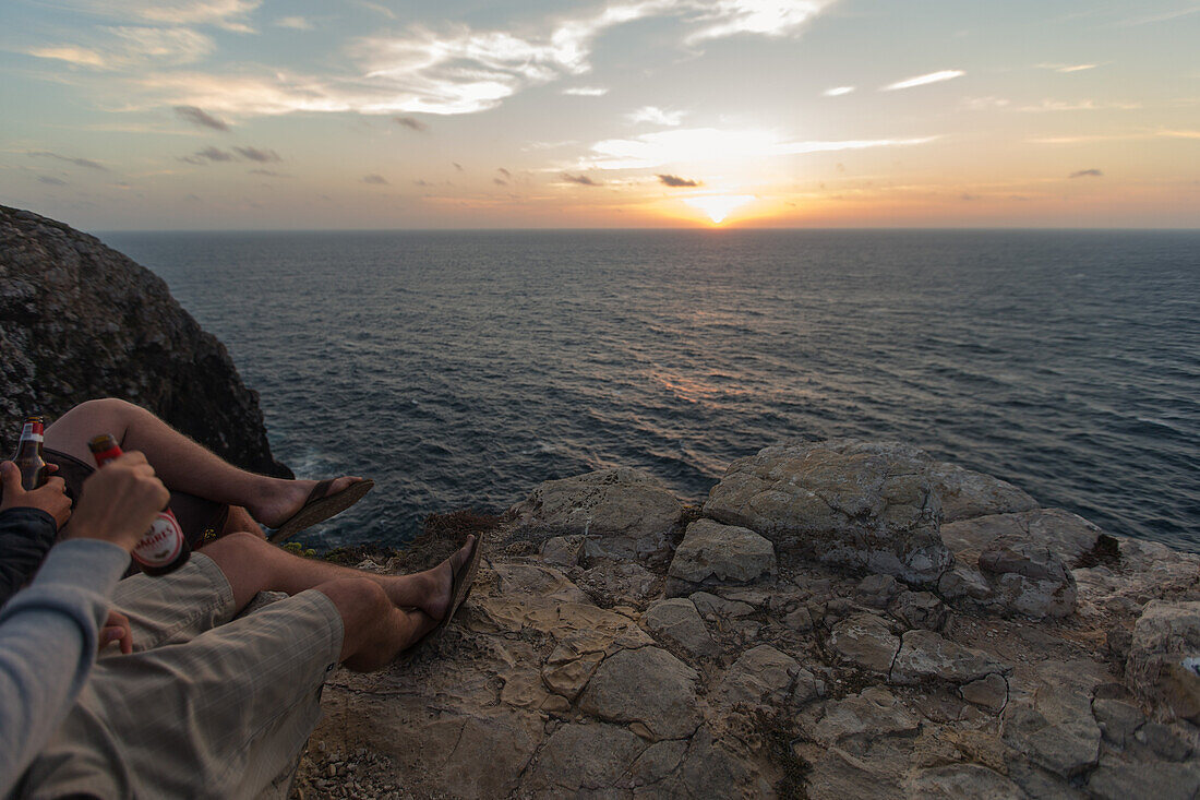 Zwei junge Männer genießen den Sonnenuntergang auf einer Klippe am Strand Praia da Amoreira,  Aljezur, Faro, Portugal