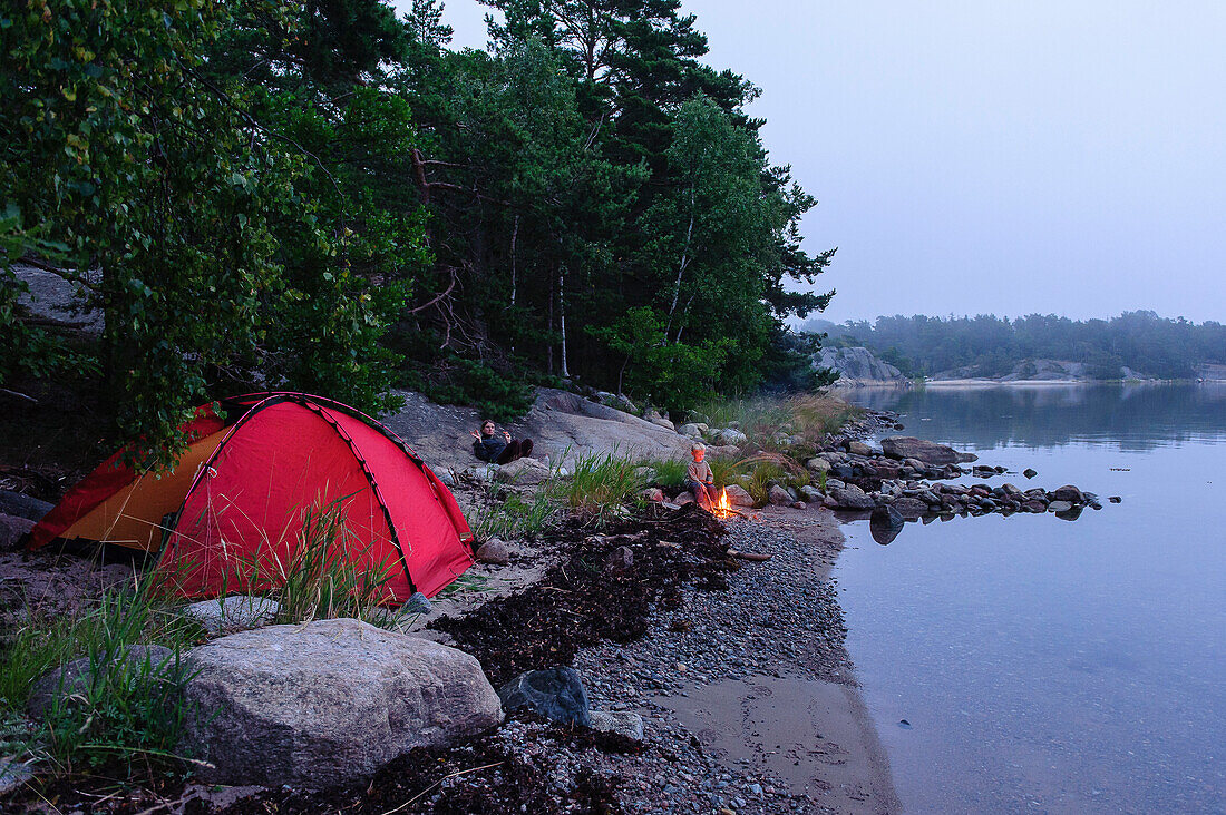 little boy next to red tent sits by the campfire in the archipelago island Fiskhamn, Stockholm, Sweden