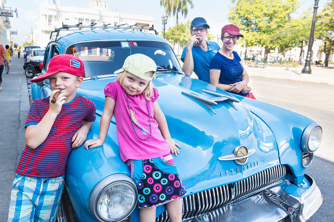 family standing in front of an blue oldtimer, pretending to smoke a cigar, at park Parque Jose Marti in the center of Cienfuegos, colonial town, family travel to Cuba, parental leave, holiday, time-out, adventure, MR, Cienfuegos, Cuba, Caribbean island
