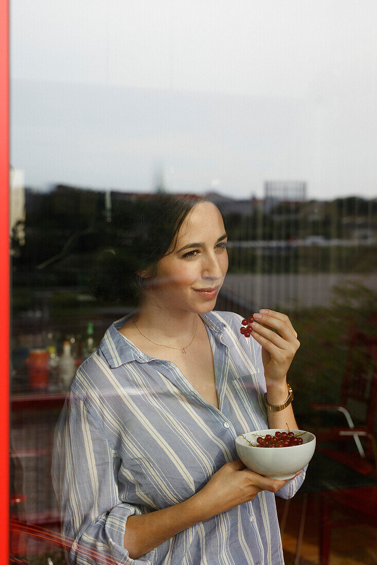 Thoughtful young woman eating red currants while looking through window
