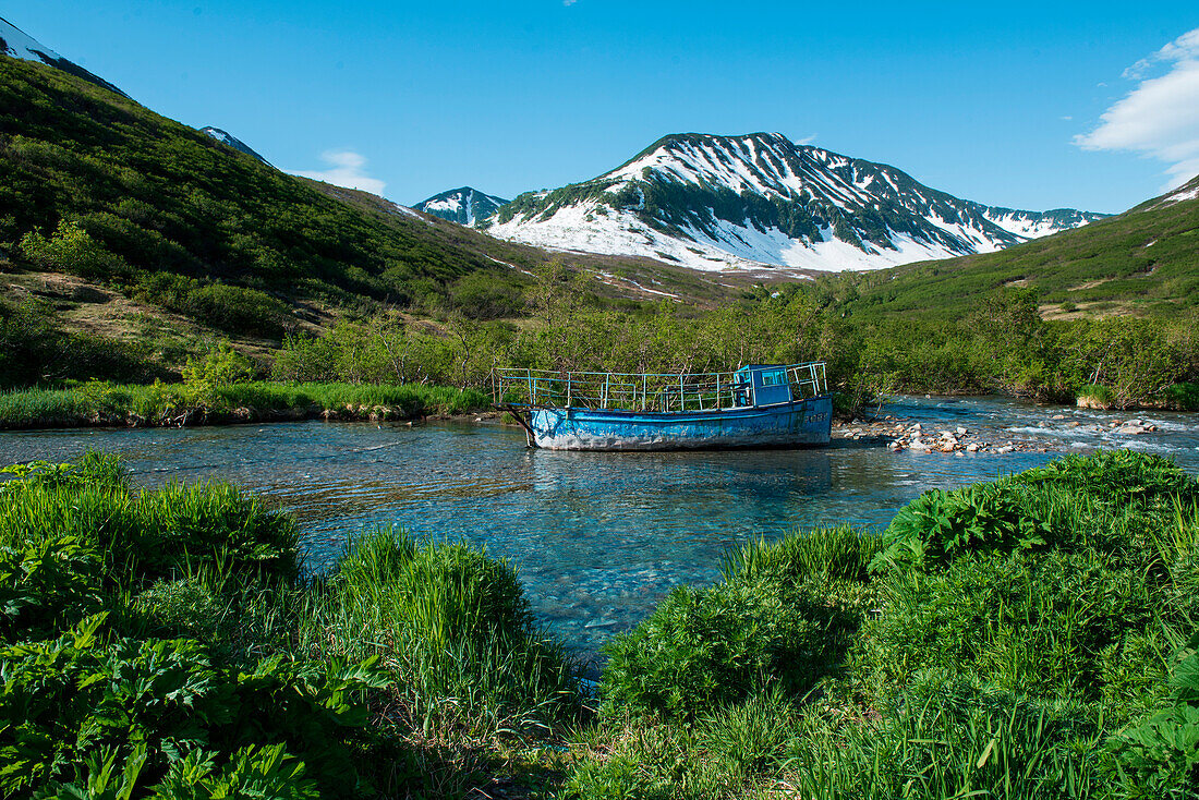 Ein heruntergekommenes blaues Boot liegt in einem Bach umgeben von Gras und Büschen mit einem Berg im Hintergrund, Russkaya Bay, Kamtschatka-Halbinsel, Russland, Asien