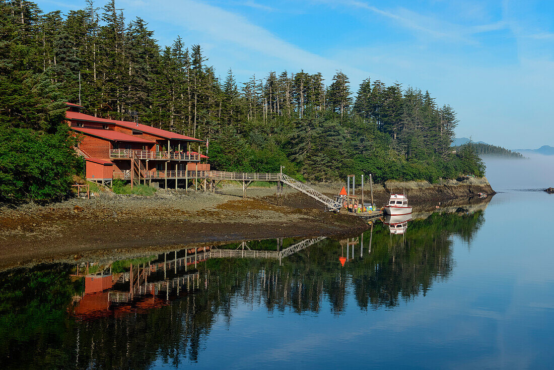 Calm weather creates a near-perfect mirror image of a large house, a pier, and a small boat on a forested peninsula, Elfin Cove, Chichagof Island, Alaska, USA, North America