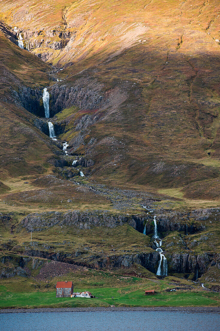 Farm buildings are dwarfed by a multiple waterfall in the steep landscape bordering Seyðisfjörðdur fjord, near Seyðisfjörðdur, Eastern Iceland, Europe