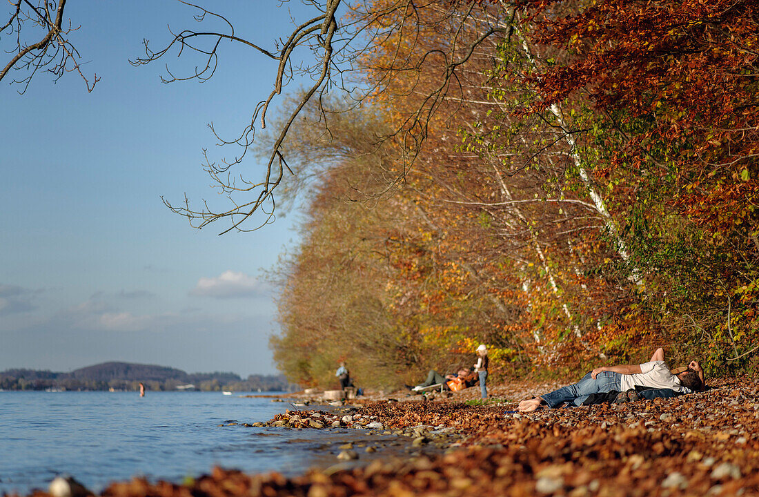 Herbst in Leoni am Starnberger See, ein Paar liegt am Ufer und geniesst die Nachmittagssonne, im Hintergrund weitere Besucher und ein Badegast, Leoni, Oberbayern, Deutschland