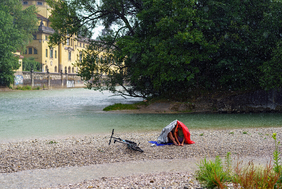 Two man protecting themselves against the rain whilst sitting on a gravel bank in the river Isar, in the background you can see the Muellersche Volksbad, Munich, Upper Bavaria, Germany