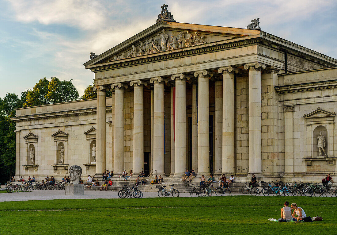 Two young ladys sitting on the grass at Koenigsplatz in front of the Ionische Glyptothek, where further people are relaxing and enjoying the evening, Munich, Upper Bavaria, Germany