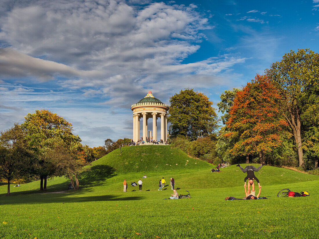 A young couple practicing yoga on the grass in front of the Monopteros in the English Garden in autumn, passersby strolling over the green, Munich, Upper Bavaria, Germany