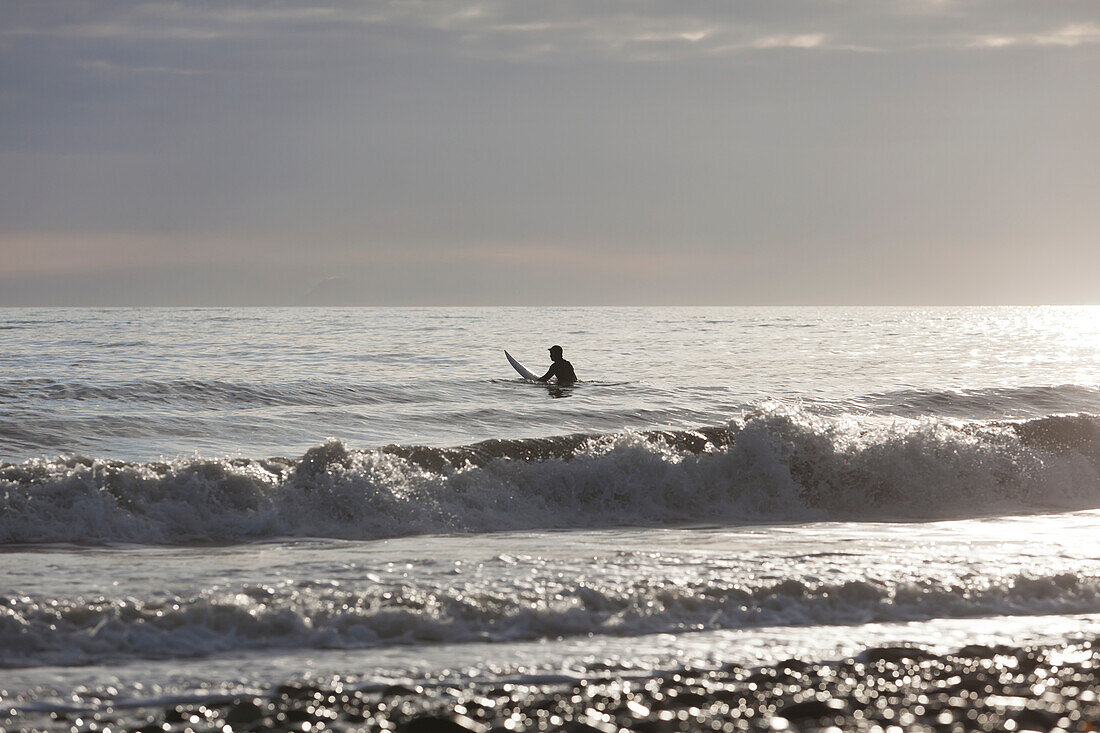 Back Lit Surfer Sitting On Surfboard In Kachemak Bay, Homer, Southcentral Alaska, USA