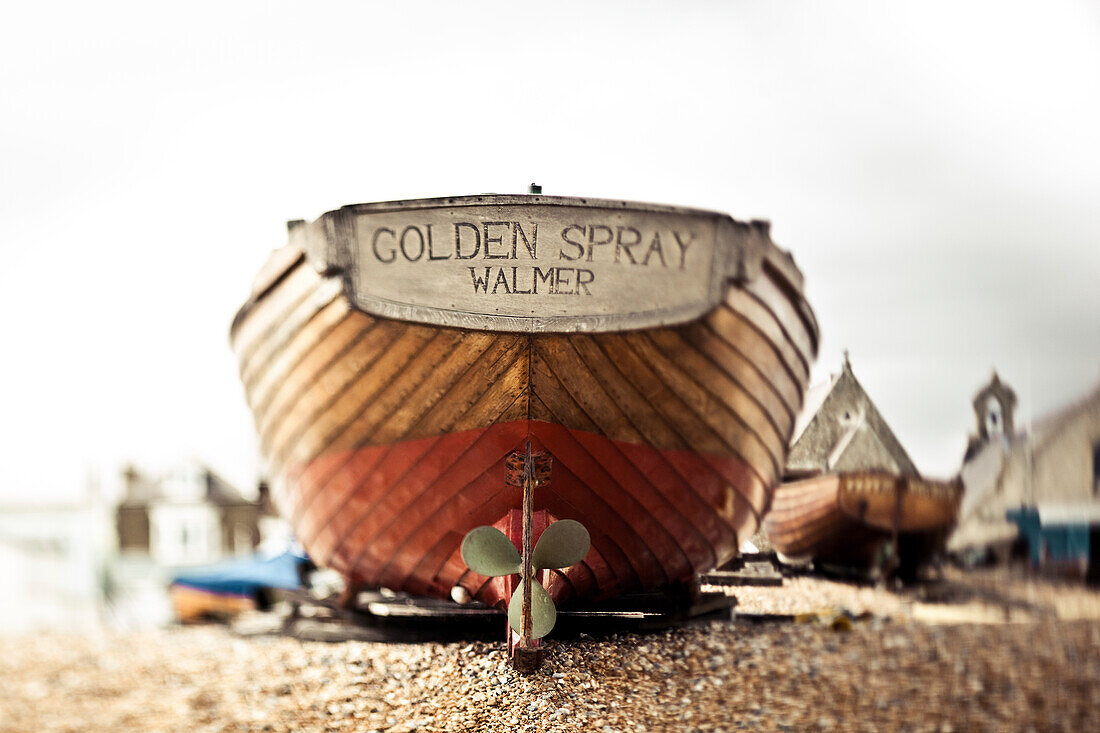 A wooden motorboat sits on the shingle beach with buildings in the background; England