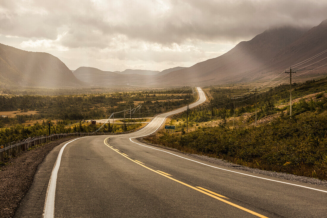 Sun rays shining through clouds over mountains and a road; Newfoundland, Canada