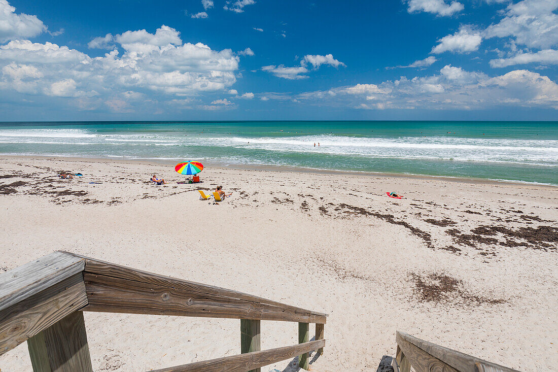 Sunbathers sit on the sand of Pelican Beach with a colourful umbrella looking out to the Atlantic ocean, and wooden steps with railing in the foreground; Satellite Beach, Florida, United States of America