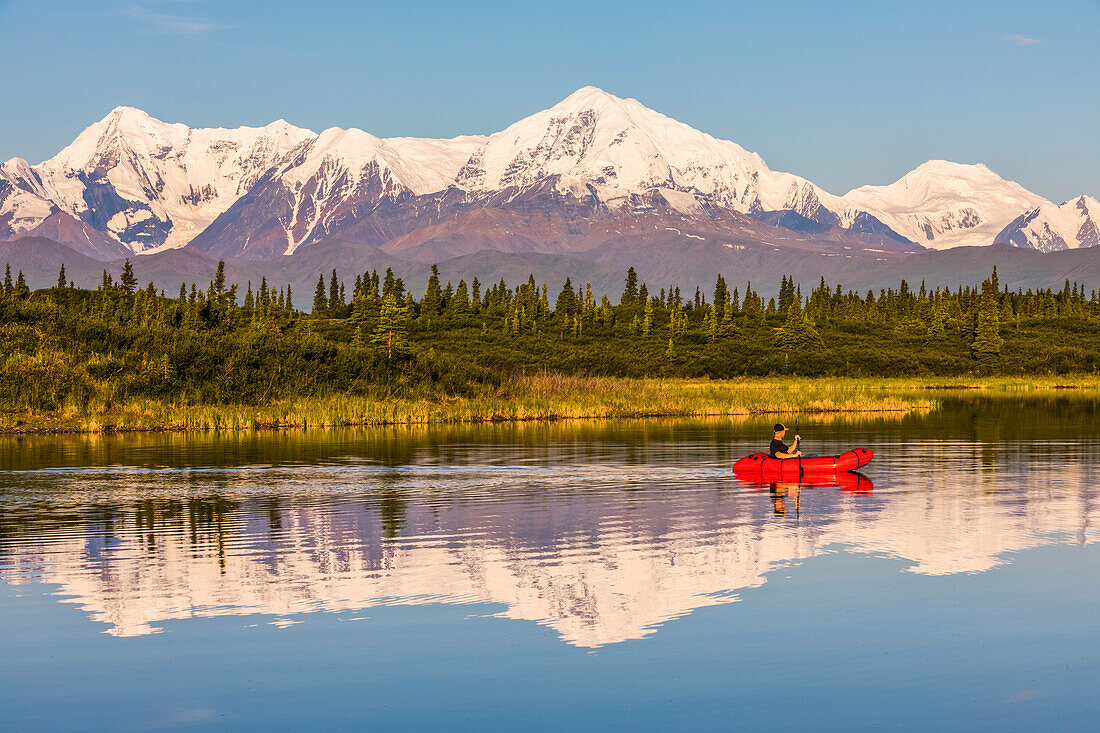 A man paddles across Donnelly Lake in a pack raft with Mt. Moffit and the Alaska Range reflecting on the water; Alaska, United States of America