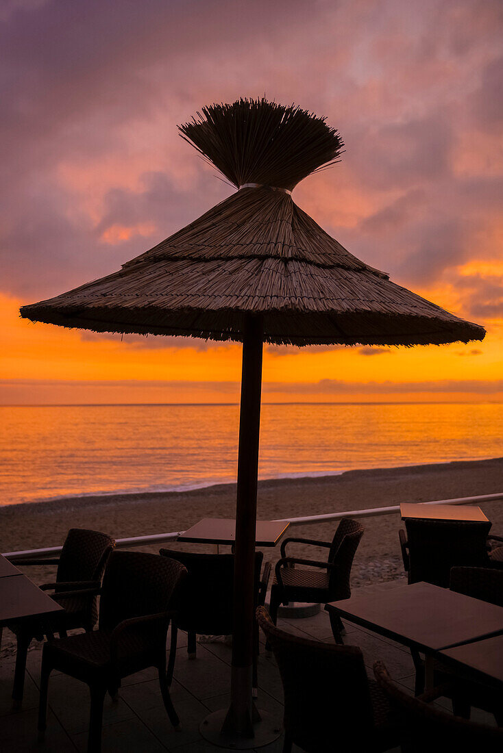 A Thatch Umbrella Over A Table With Chairs On The Beach At Sunset, Looking Out Over The Mediterranean Sea; Menton, Cote D'azur, France