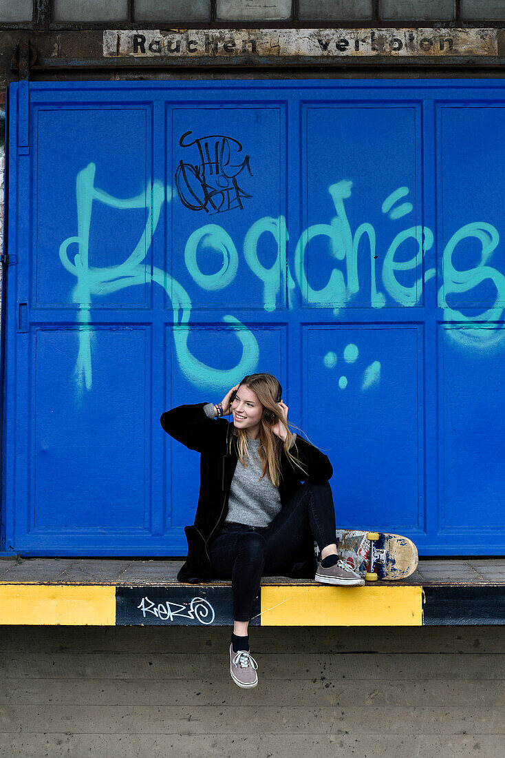 Girl with headphones and skateboard sitting in front of graffitti wall, Hamburg, Germany