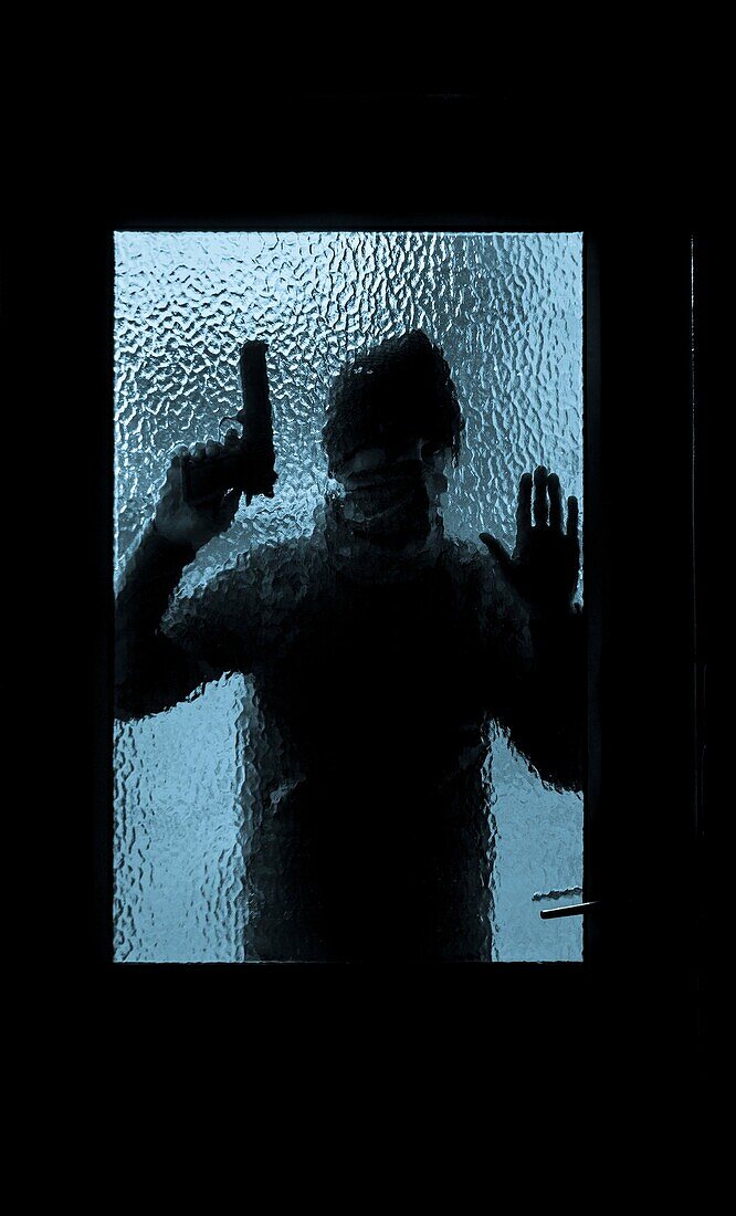 Silhouette of man holding gun looking through house window.