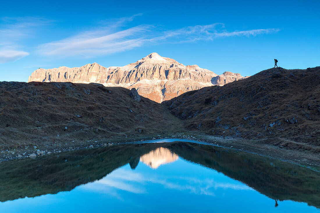 Hiker in silhouette near a small pond with Sella group with the highest Piz Boè mountain reflected, Pordoi pass, Arabba, Beuuno, Veneto, Italy