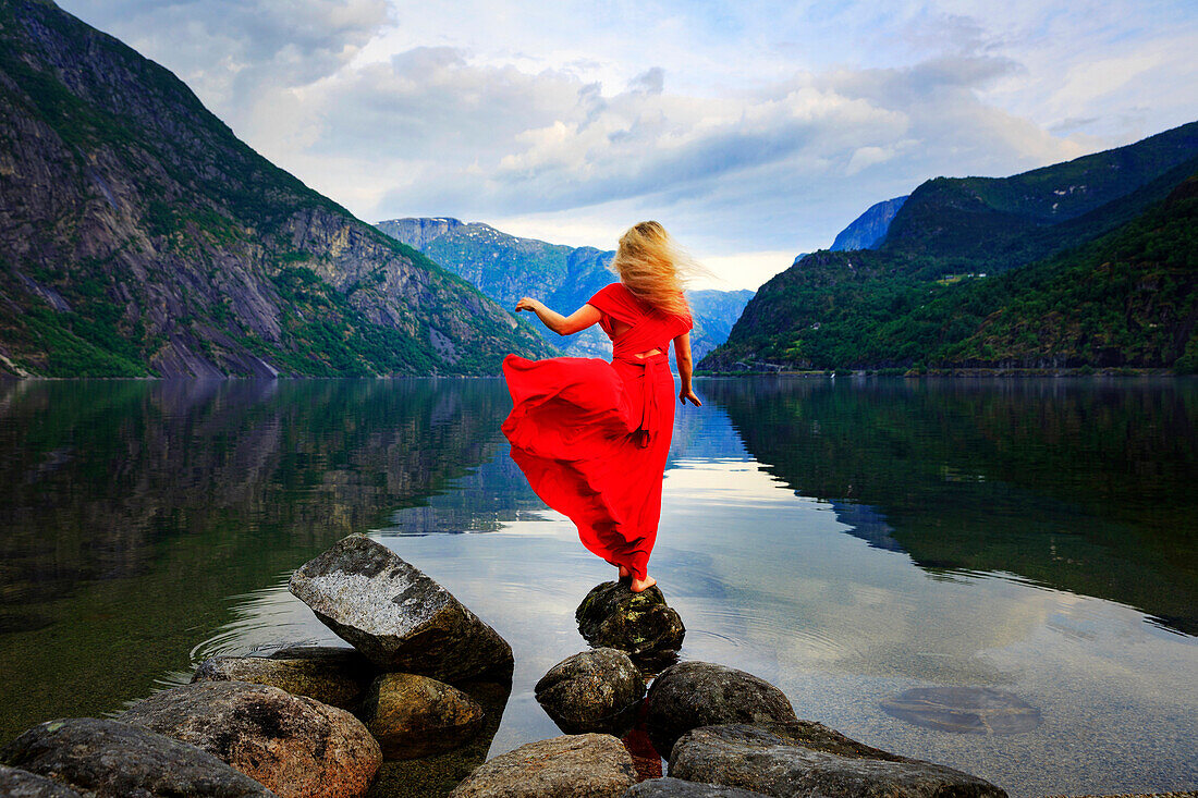 Blonde girl in a red dress moved by the wind standing on a rock in front of a lake, Eidfjord, Hordaland, Norway