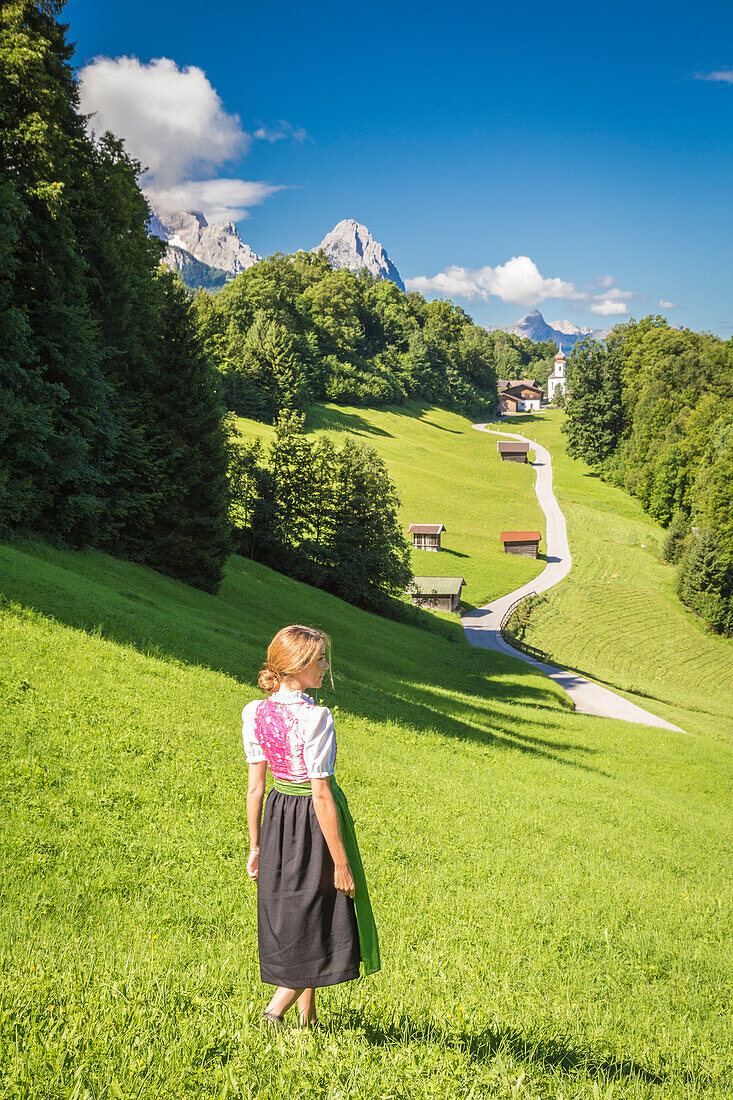 A girl in Typical Bayern dress walking in front of Wamberg village, with Mount Zugspitze and Waxenstein on the background, Garmisch Partenkirchen, Bayern, Germany