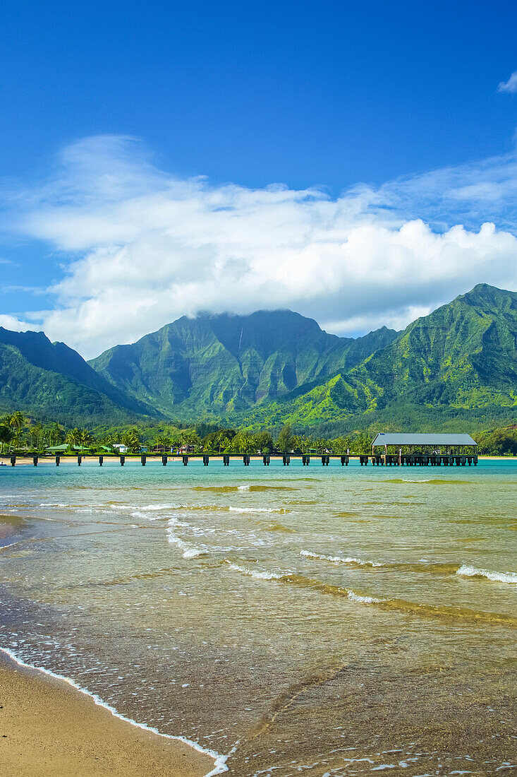 A pier leading out to the ocean water along the coastline with water washing onto the beach in the foreground and rugged, green mountains in the background; Hanalei, Kauai, Hawaii, United States of America