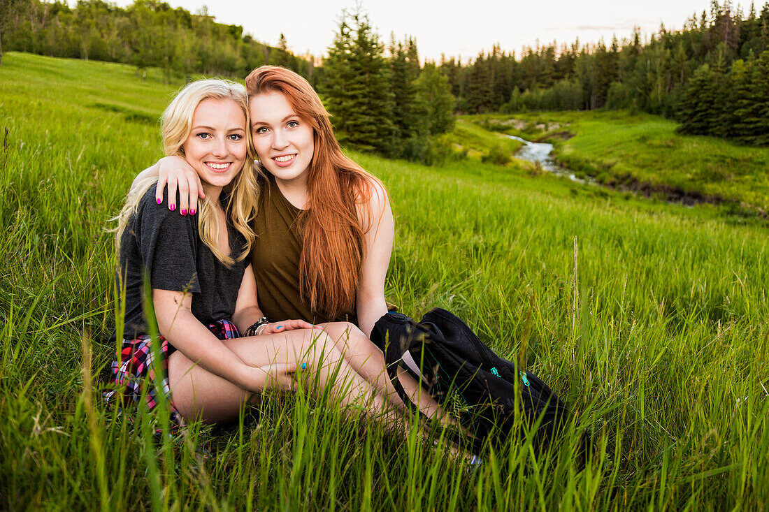 Zwei Freundinnen sitzen in einer Umarmung auf einer Wiese und posieren für ein Porträt bei Sonnenuntergang; Edmonton, Alberta, Kanada