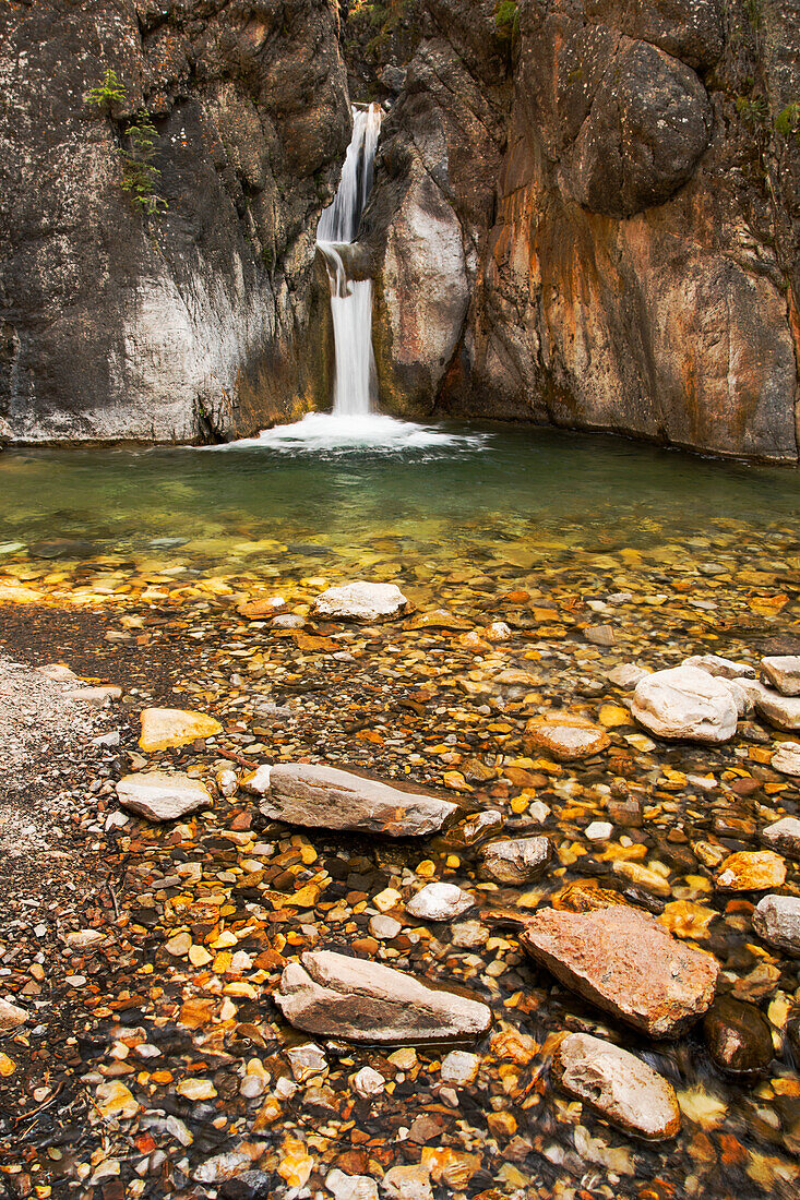 A double waterfall between dark rock walls with the foreground of colourful small rocks and pebbles; Calgary, Alberta, Canada