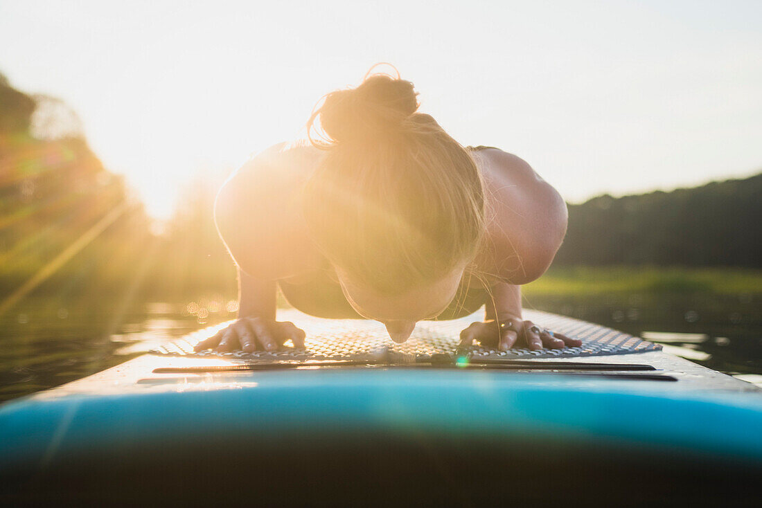 Young woman doing yoga Chaturanga, Staff Pose on stand up paddle board at golden hour, North Kingstown, Rhode Island, USA